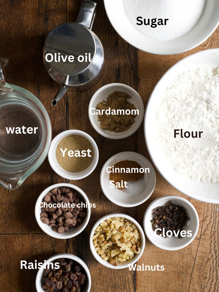 Ingredients to make lazarakia buns: Water, olive oil, cardamom, cinnamon, sugar, flour, yeast, salt, cloves, chocolate chips, raisins, walnuts
