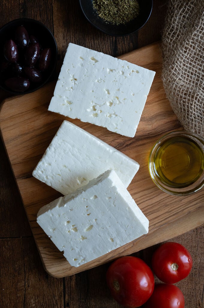 Blocks of feta cheese on a wooden board, surrounded by olive oil, olives, and oregano.