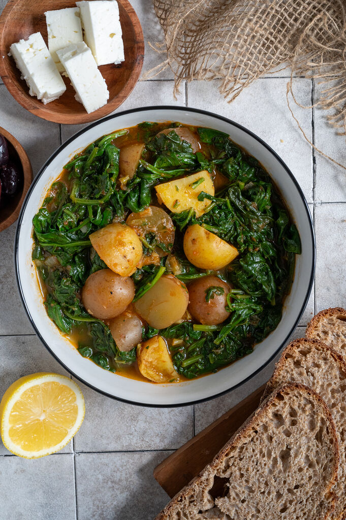 Spinach and potato stew in a white bowl, surrounded by feta, olives and bread.
