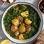 Spinach and potato stew in a white bowl, surrounded by feta, olives and bread.