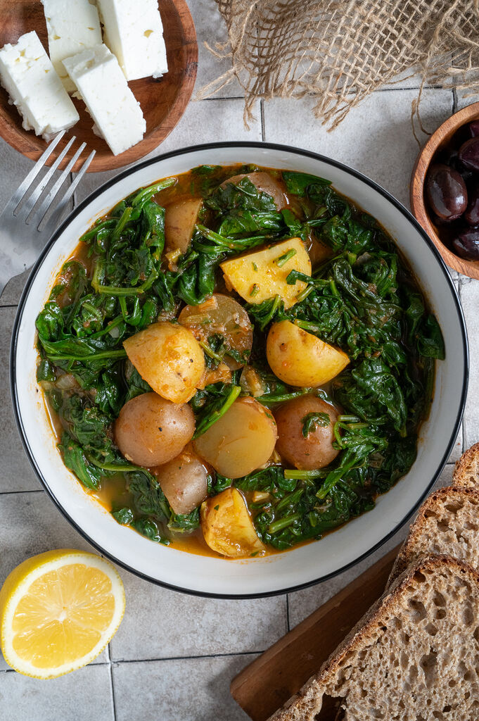 Spinach and potato stew in a white bowl, surrounded by feta, olives and bread.
