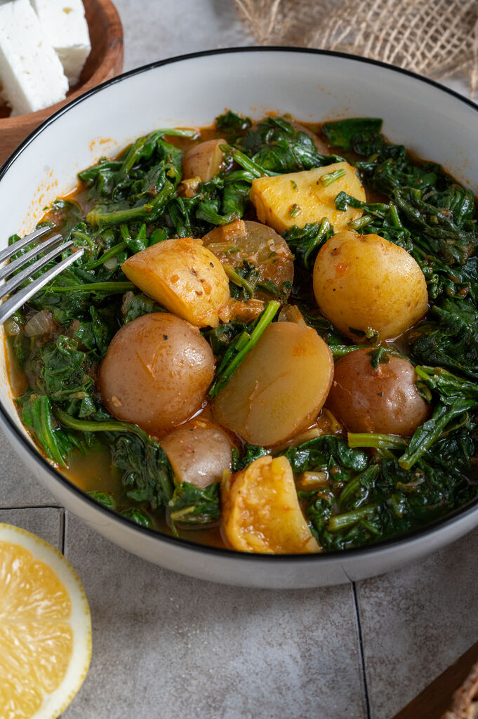Spinach and potato stew in a white bowl, surrounded by feta, olives and bread.