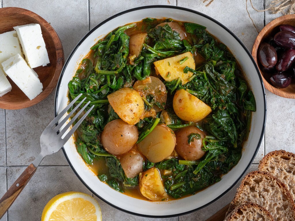 Spinach and potato stew in a white bowl, surrounded by feta, olives and bread.