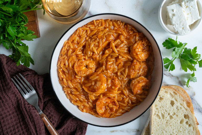 Garides youvetsi, Greek shrimp with orzo. In a white bowl surrounded by feta, bread, a fork, a napkin and wine.