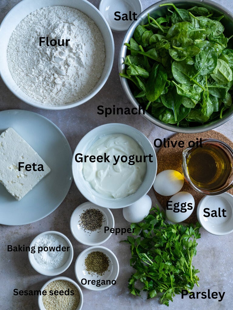 Ingredients such as flour, salt, spinach, feta, greek yoghourt, olive oil, eggs, Pepper, baking powder, oregano, parsley and sesame seeds.
