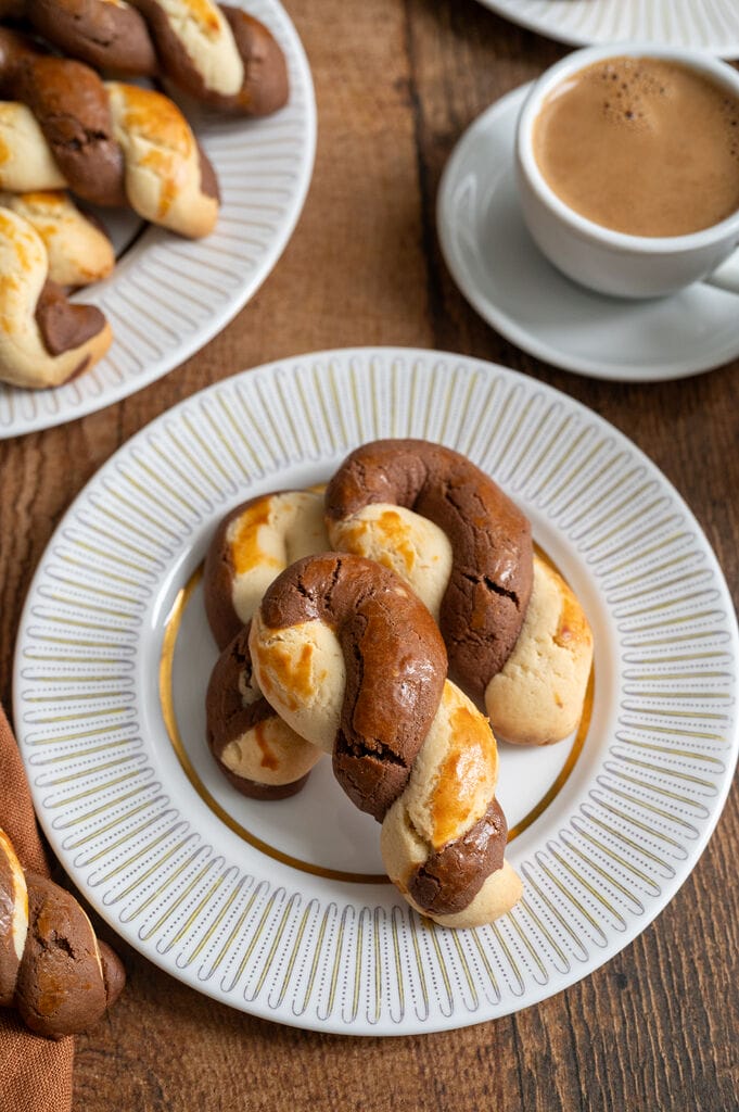 Greek butter koulourakia with a chocolate twist on a white place with gold decor, and a cup of Greek coffee next to it.