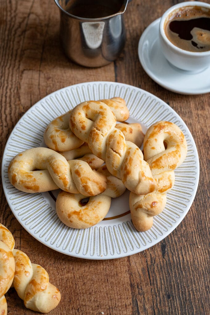 Greek butter koulourakia on a white plate with gold decor, next to a cup of Greek coffee.