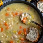 Fasolatha Greek soup in a gray bowl, surrounded by bread and olives and on a gray/blue backdrop.