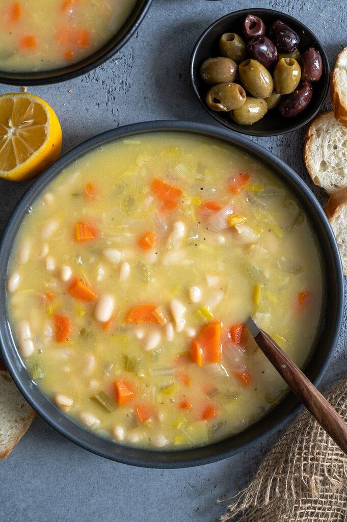 Fasolatha Greek soup in a gray bowl, surrounded by bread and olives and on a gray/blue backdrop.