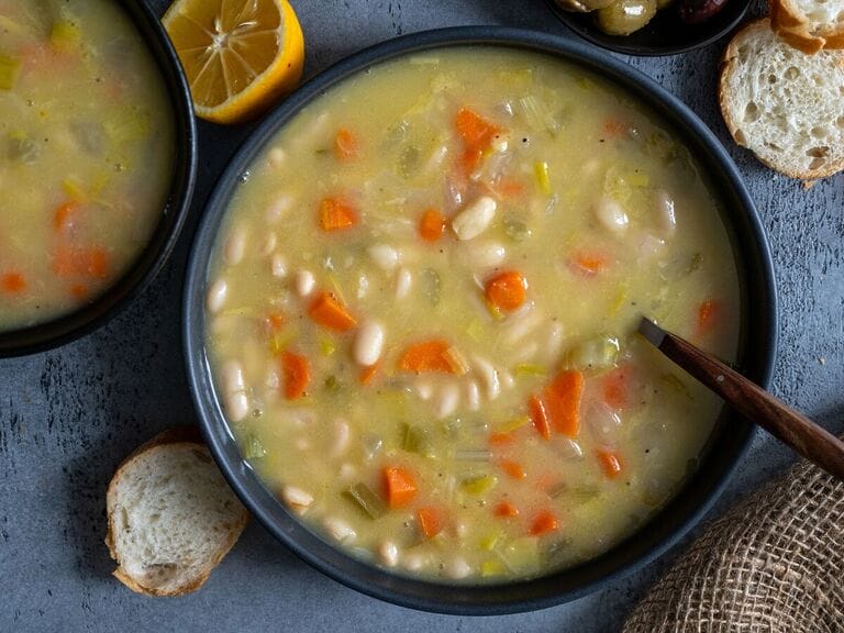 Fasolatha Greek soup in a gray bowl, surrounded by bread and olives and on a gray/blue backdrop.