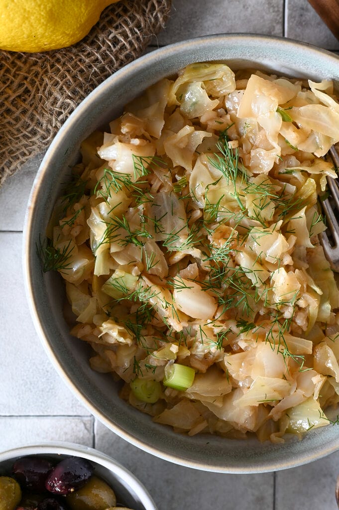 Lahanorizo, a Greek cabbage and rice meal plated in a bowl surrounded by lemons, bread and olives, and sprinkled with some dill.