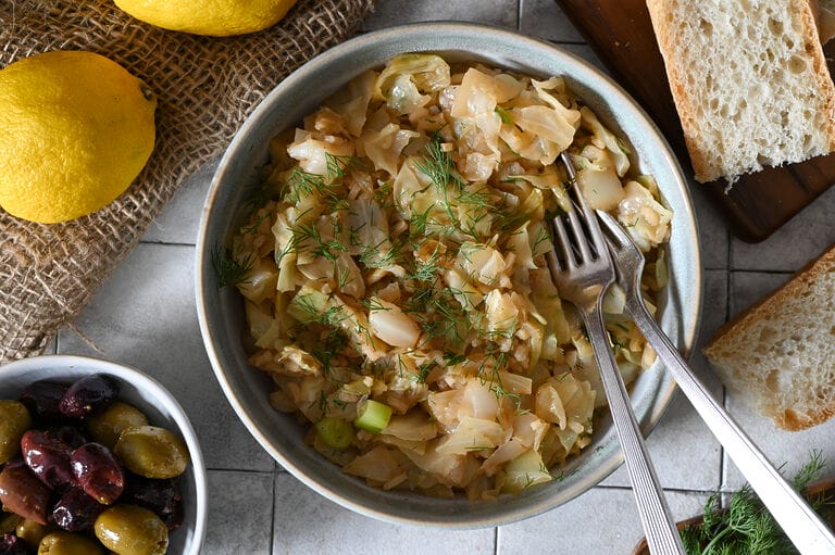 Lahanorizo, a Greek cabbage and rice meal plated in a bowl surrounded by lemons, bread and olives, and sprinkled with some dill.