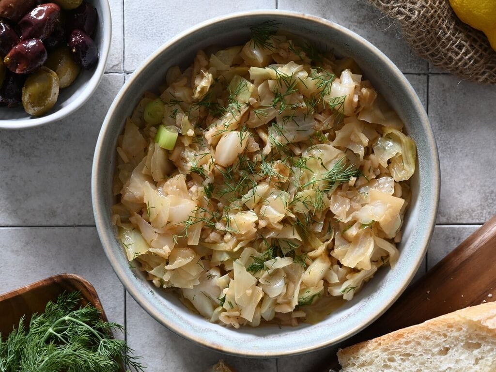 Lahanorizo, a Greek cabbage and rice meal plated in a bowl surrounded by lemons, bread and olives, and sprinkled with some dill.