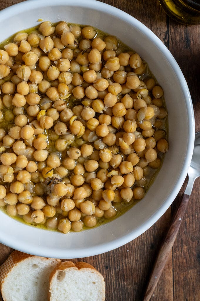 Baked chickpeas in a white bowl, surrounded by bread, and olives.