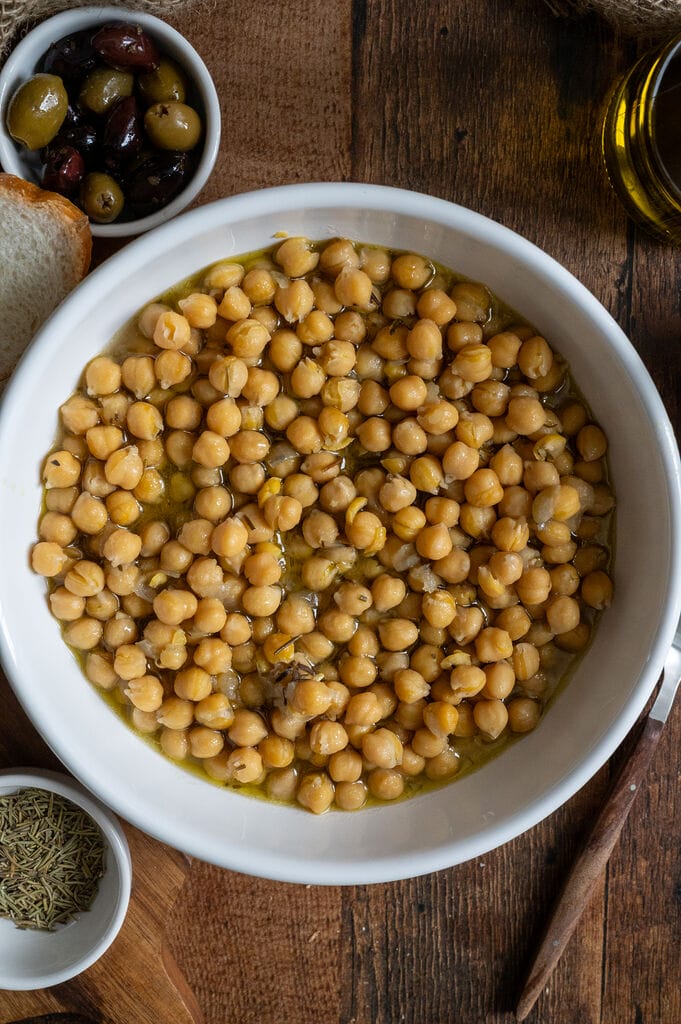 Baked chickpeas in a white bowl, surrounded by bread, and olives.