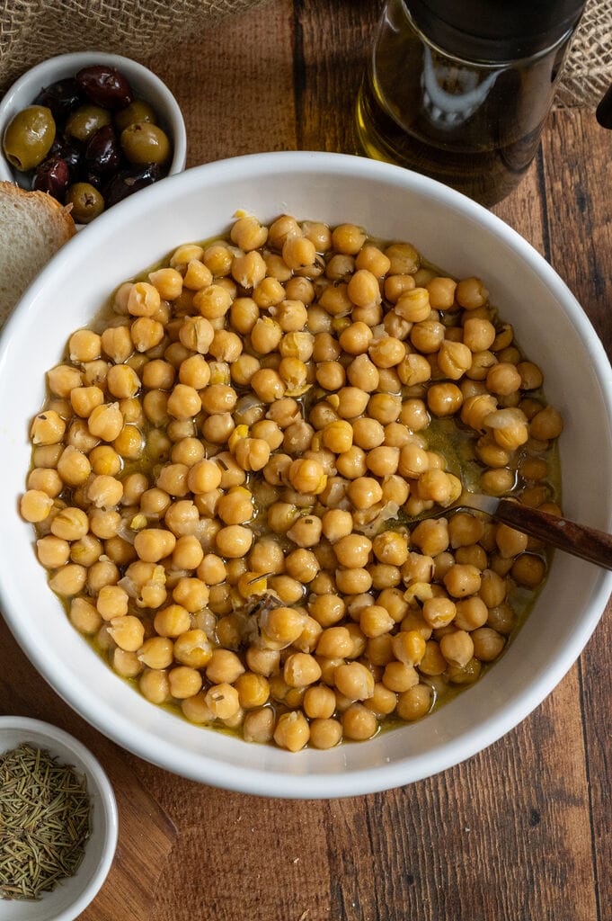Baked chickpeas in a white bowl, surrounded by bread, and olives.