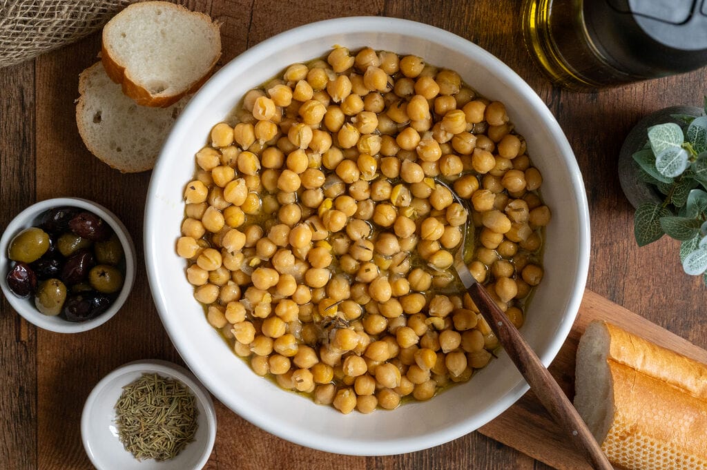 Baked chickpeas in a white bowl, surrounded by bread, and olives.