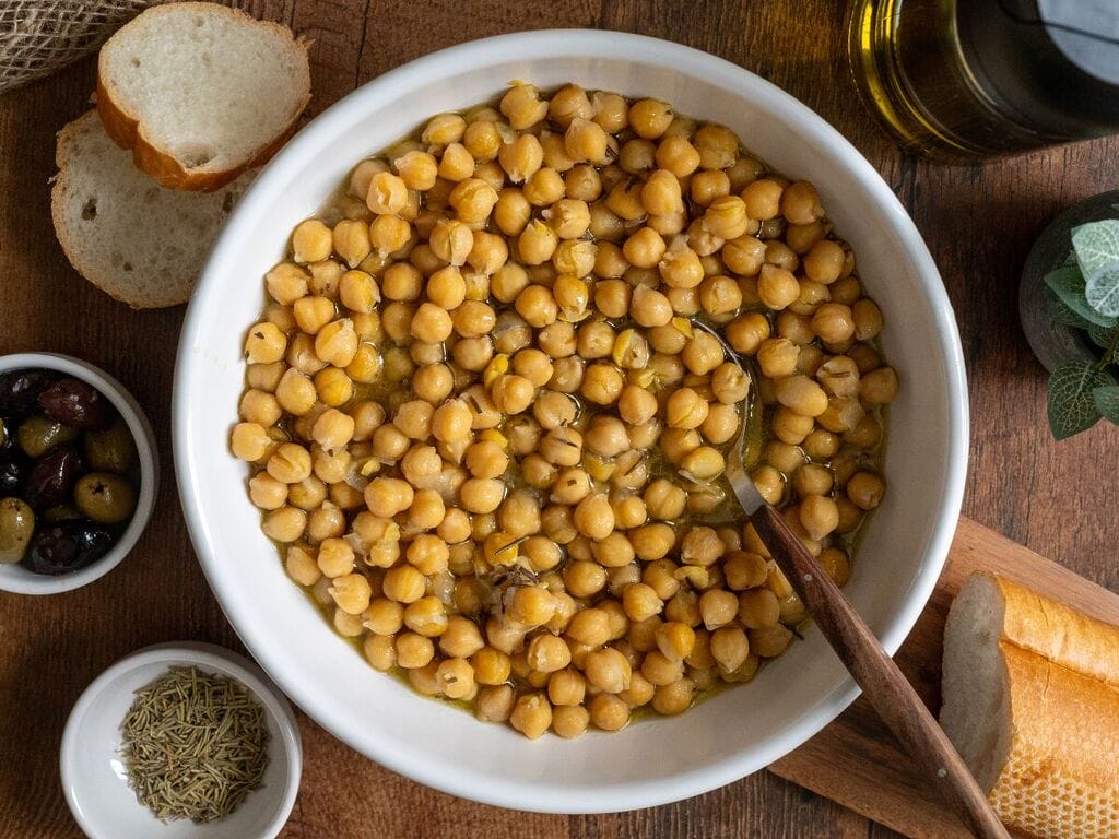 Baked chickpeas in a white bowl, surrounded by bread, and olives.