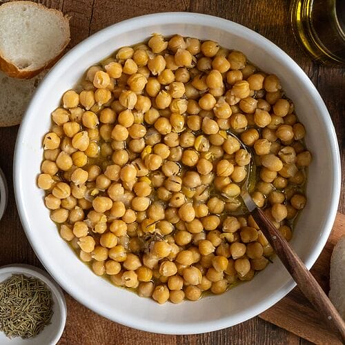 Baked chickpeas in a white bowl, surrounded by bread, and olives.