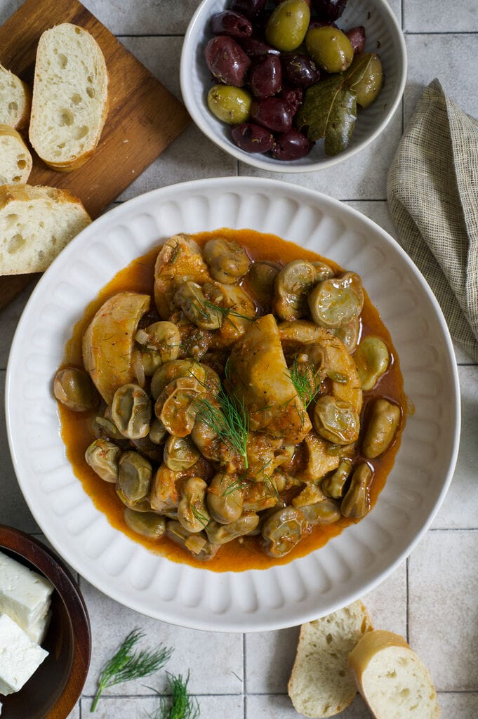 Greek artichokes with fava beans (Aginares me koukia) on a white plate surrounded by bread, olives and feta.