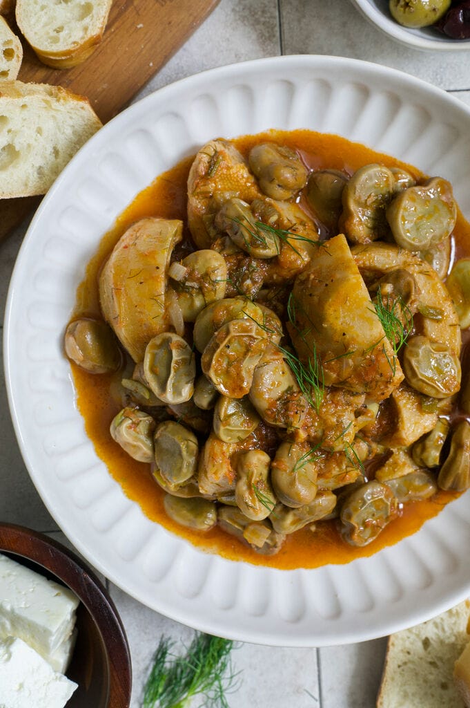 Greek artichokes with fava beans (Aginares me koukia) on a white plate surrounded by bread, olives and feta.