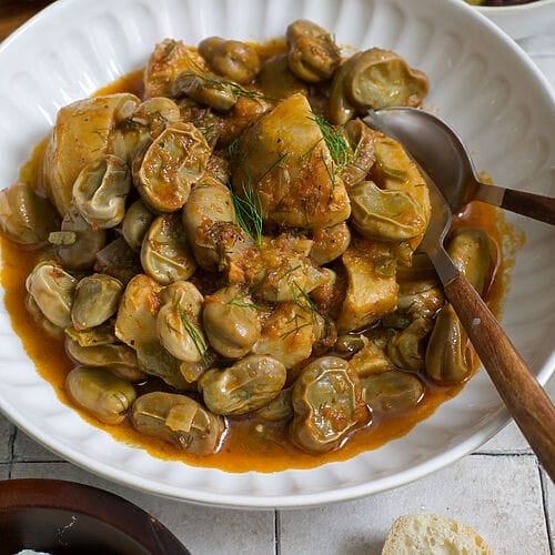 Greek artichokes with fava beans (Aginares me koukia) on a white plate surrounded by bread, olives and feta.