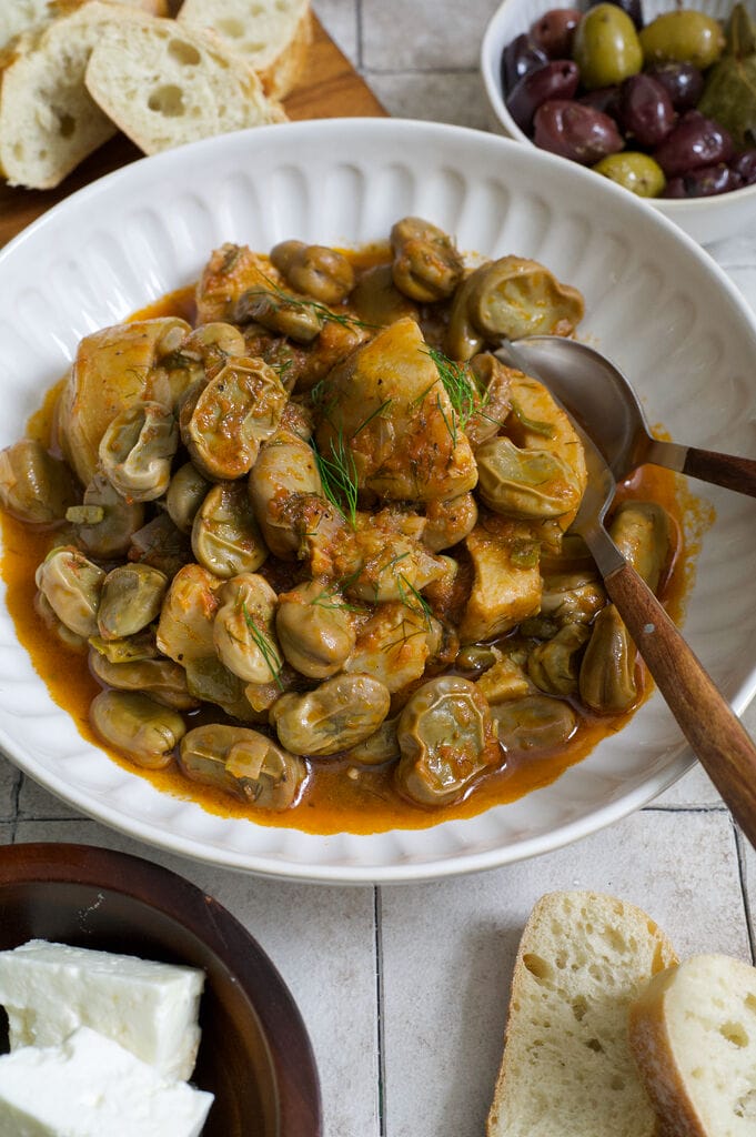 Greek artichokes with fava beans (Aginares me koukia) on a white plate surrounded by bread, olives and feta.