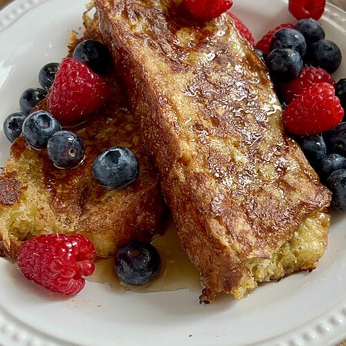 Tsoureki French toast, Greek Easter bread French toast on a white plate, surrounded by berries and topped with maple syrup.