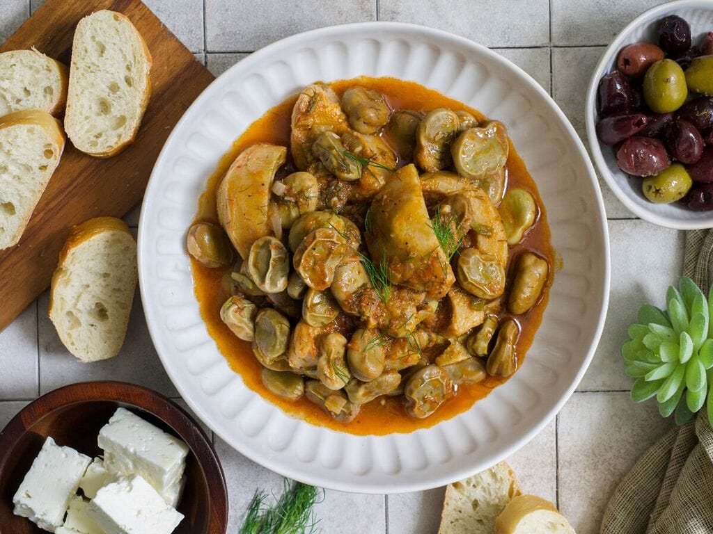 Greek artichokes with fava beans (Aginares me koukia) on a white plate surrounded by bread, olives and feta.