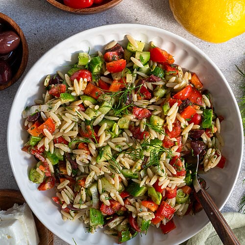 Greek pasta salad in a white bowl, surrounded by feta, olives, and a lemon.