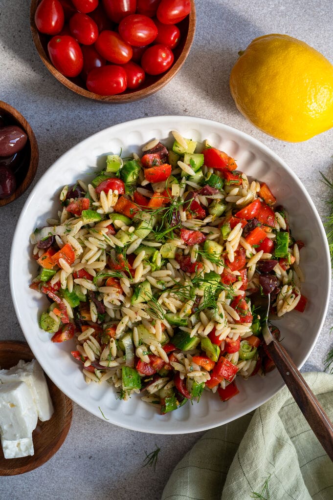 Greek pasta salad in a white bowl, surrounded by feta, olives, and a lemon.