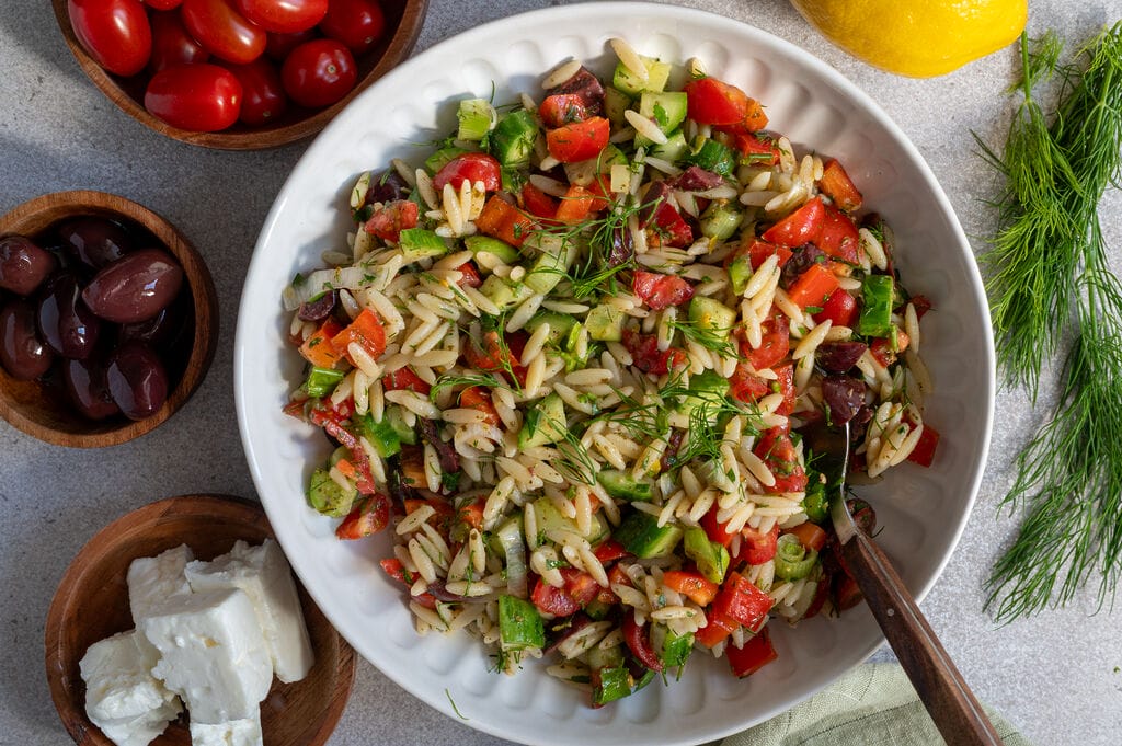 Greek pasta salad in a white bowl, surrounded by feta, olives, and a lemon.