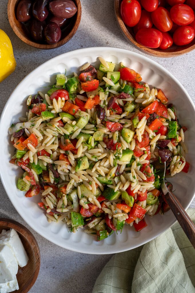 Greek pasta salad in a white bowl, surrounded by feta, olives, and a lemon.