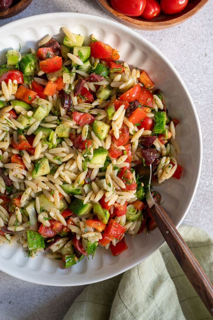 Greek pasta salad in a white bowl, surrounded by feta, olives, and a lemon.