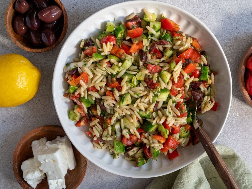 Greek pasta salad in a white bowl, surrounded by feta, olives, and a lemon.