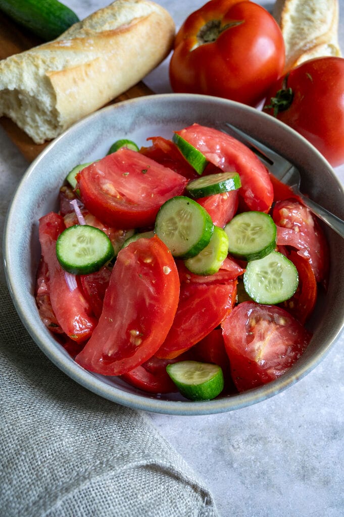 Tomato and cucumber salad in a bowl, surrounded by tomatoes, and bread.