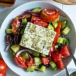 Greek salad in a white bowl, topped with feta, surrounded by bread.