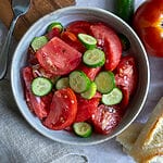 Tomato and cucumber salad in a bowl, surrounded by tomatoes, and bread.