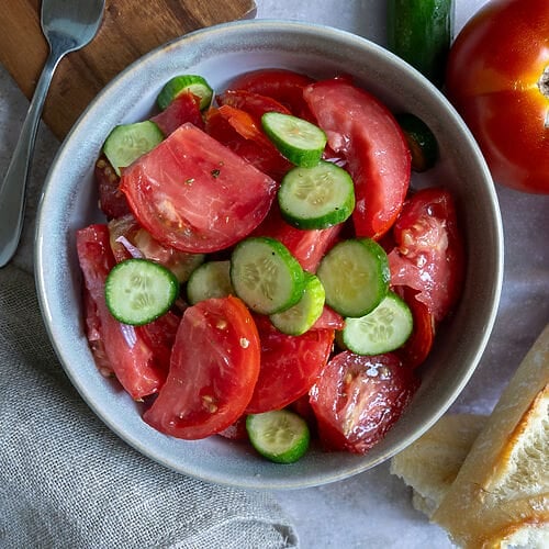 Tomato and cucumber salad in a bowl, surrounded by tomatoes, and bread.