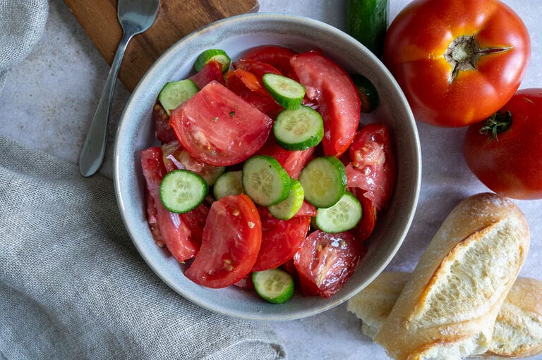 Tomato and cucumber salad in a bowl, surrounded by tomatoes, and bread.