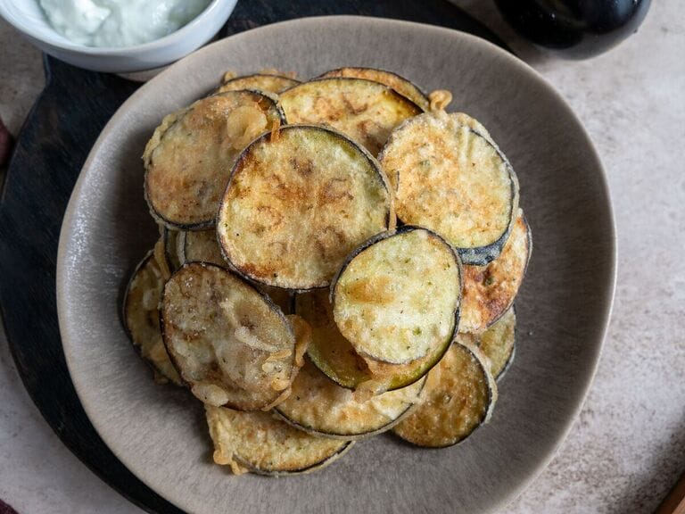 Fried eggplant chips on a brown plate surrounded by tzatziki.