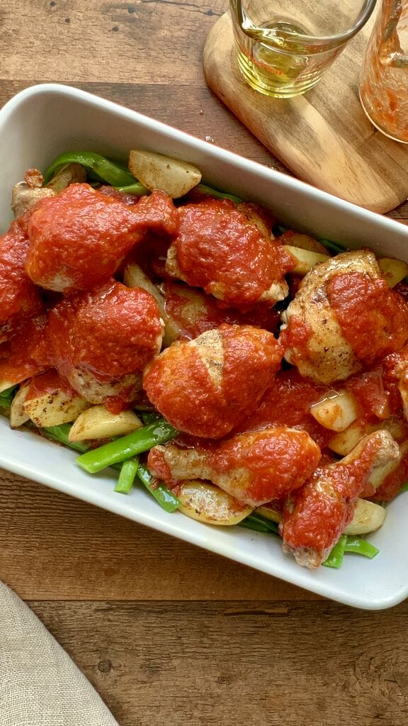 The green beans and potatoes and chicken topped with tomato sauce in a white casserole dish.