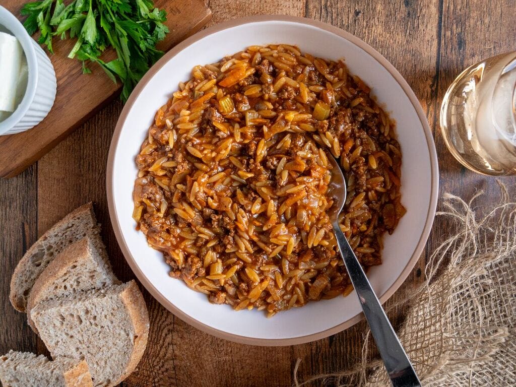 Greek orzo with ground beef, Youvetsi me kima stin katsarola, in a white bowl surrounded by bread, feta, parsley and water.
