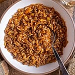 Greek orzo with ground beef, Youvetsi me kima stin katsarola, in a white bowl surrounded by bread, feta, parsley and water.