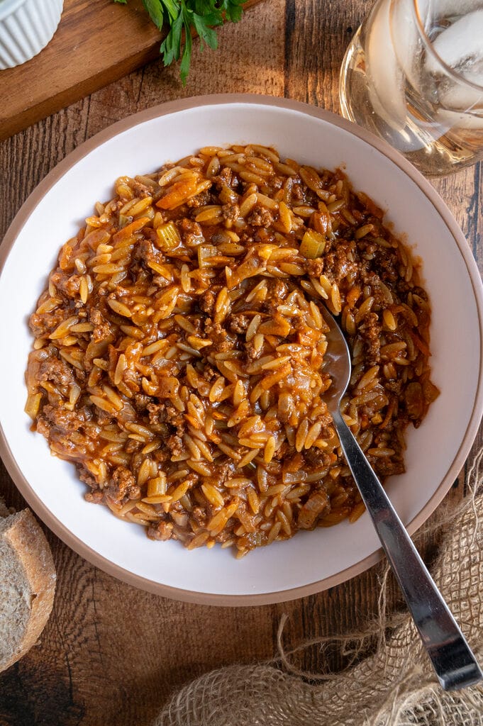Greek orzo with ground beef, Youvetsi me kima stin katsarola, in a white bowl surrounded by bread, feta, parsley and water.
