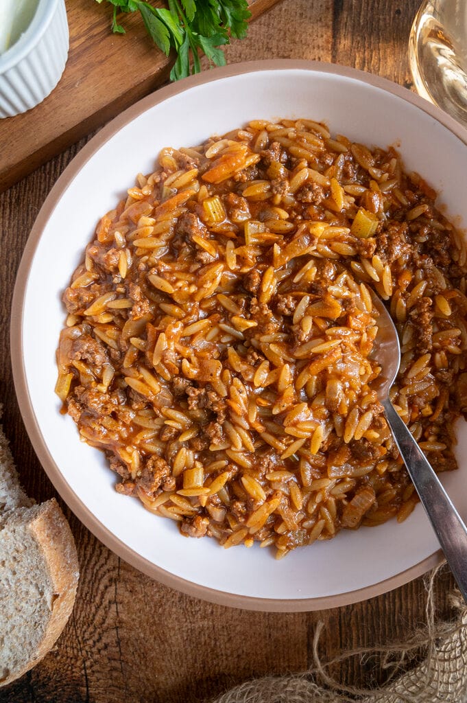 Greek orzo with ground beef, Youvetsi me kima stin katsarola, in a white bowl surrounded by bread, feta, parsley and water.