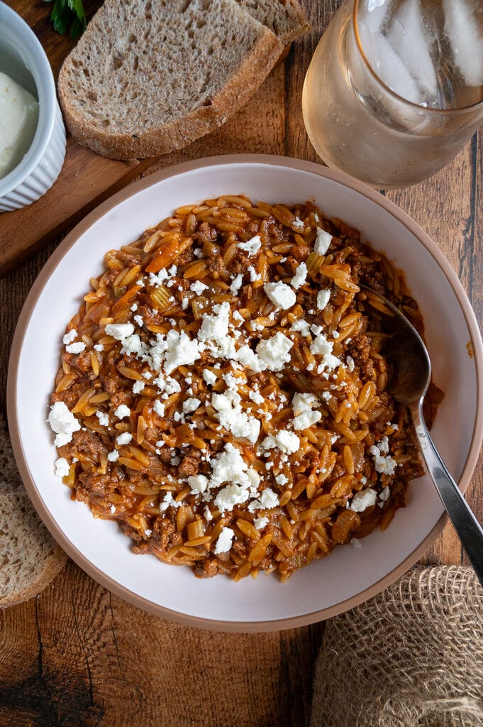 Greek orzo with ground beef, Youvetsi me kima stin katsarola, in a white bowl surrounded by bread, feta, parsley and water. Topped with feta.
