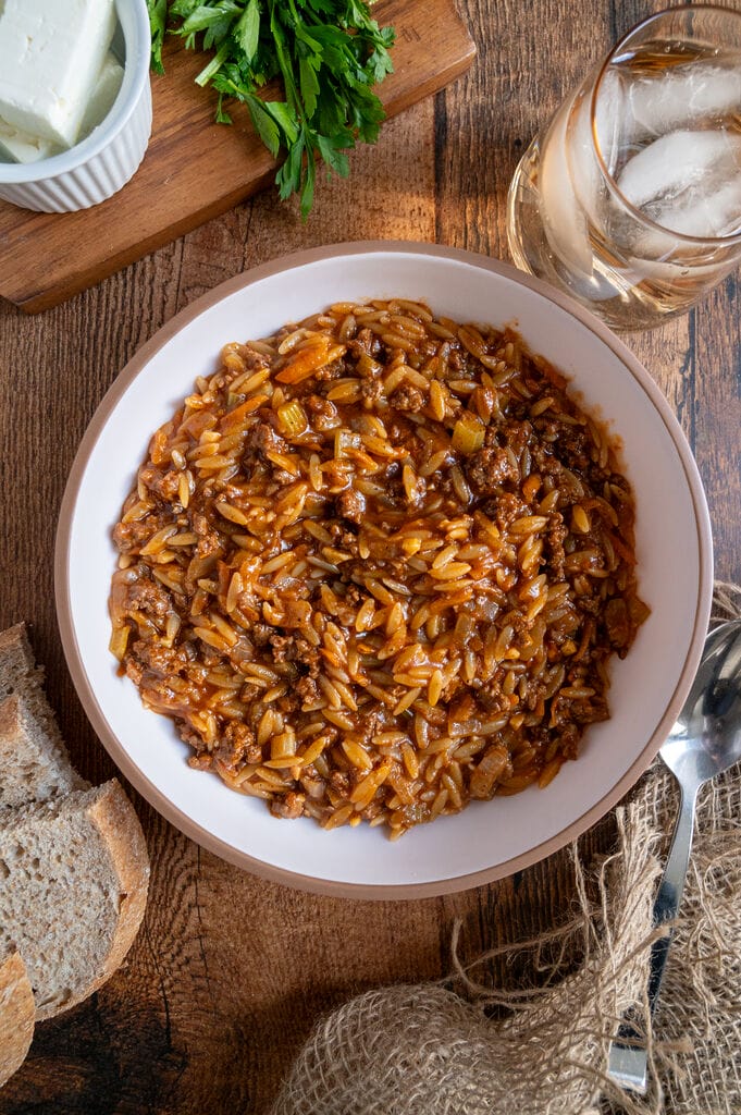 Greek orzo with ground beef, Youvetsi me kima stin katsarola, in a white bowl surrounded by bread, feta, parsley and water.