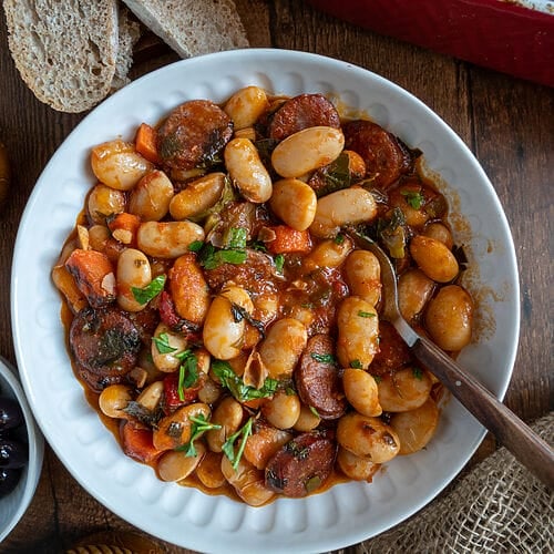 Greek Baked Gigantes with Sausage (Loukaniko) in a white bowl, surrounded by bread, and olives, and feta.