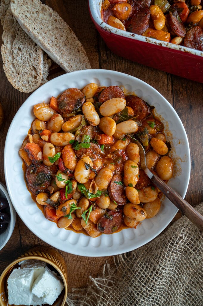 Greek Baked Gigantes with Sausage (Loukaniko) in a white bowl, surrounded by bread, and olives, and feta.